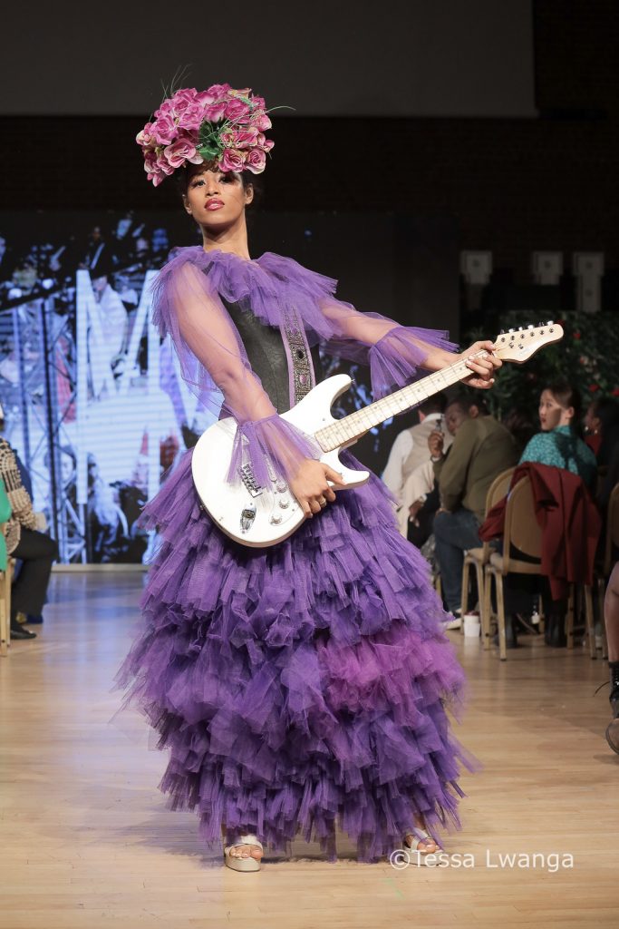Model wears purple taffeta layered ball gown by Mary Martin London whilst playing guitar on catwalk