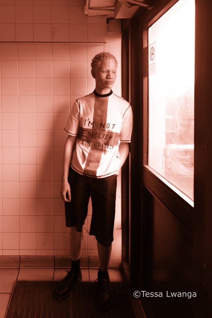 LFWAW25-0698 Albino model leans against doorframe, wearing statement punk t-shirt.