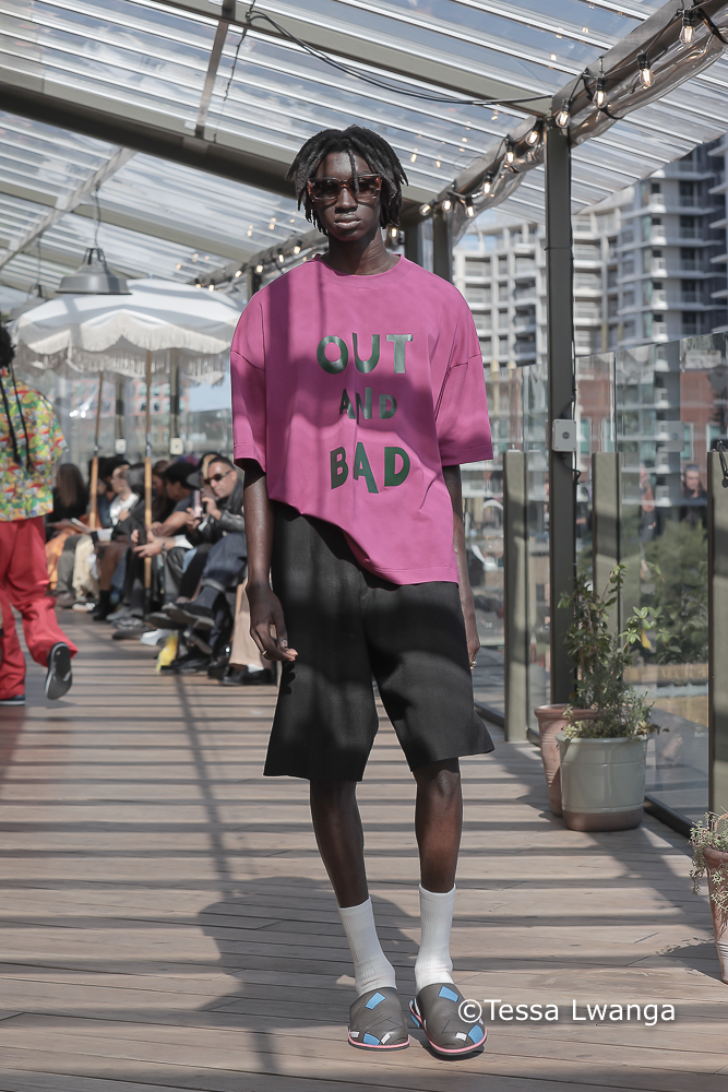 Sagaboi-0397 Model wears purple T-Shirt with inscription 'Out and Bad',on Sagaboi London Fashion Catwalkon a partially sunny and shaded terrace