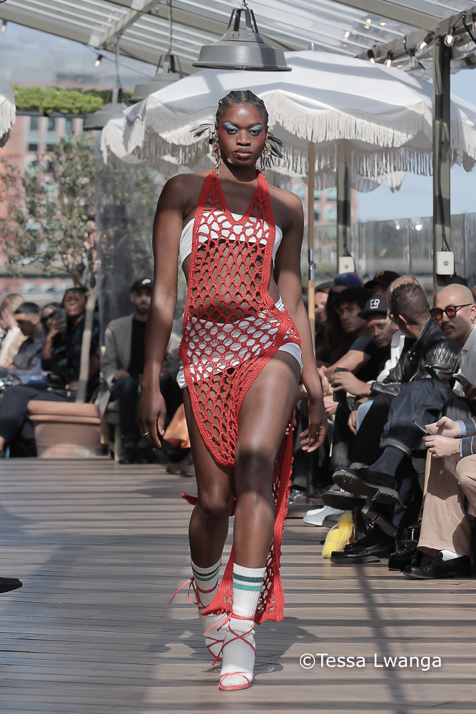 Sagaboi-0508 Model wears bright red crotchet dress over a white top and skirt at Sagaboi London Fashion Week Catwalk Show on a partially shaded and sunny terrace in White City, London