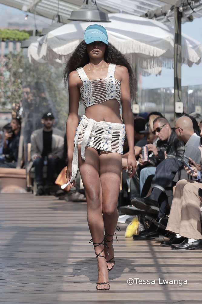 Sagaboi-0687 Model with branded Sagaboi baseball cap, long curly hair and white crotchet top and skirt on London Fashion Week Catwalk for Sagaboi SS25 Show.
