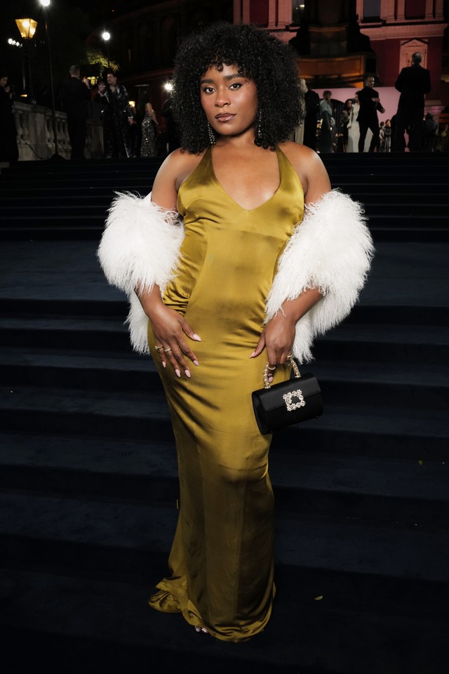 Magazine Editor, wears curly hairstyle with golden gown and furry stole