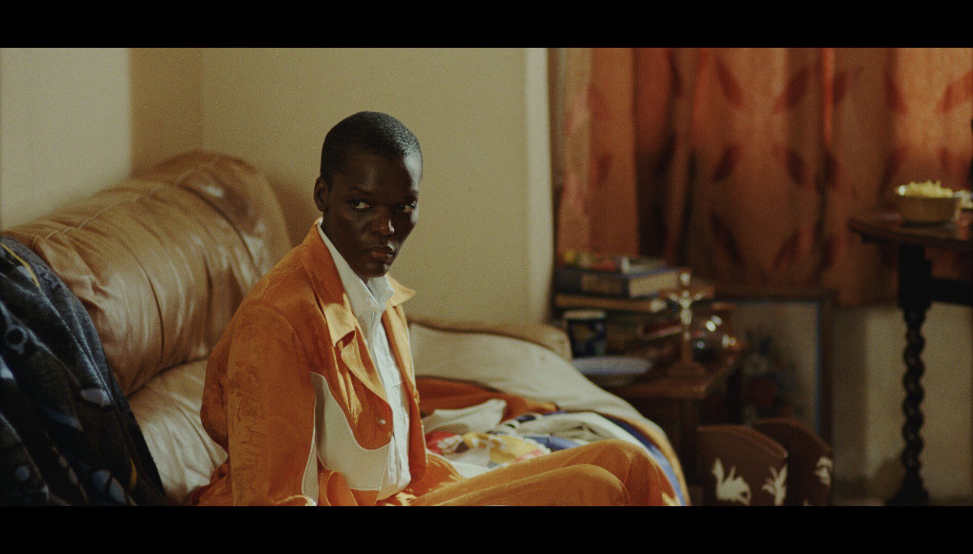 Photograph of Black Actress, Sheila Atim, wearing an orange outfit, sat on a sofa looking to the left with a serious face