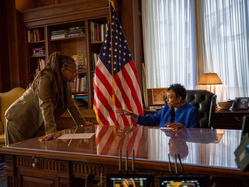 Director Ava DuVernay stands at desk with an elderly woman sat down and American Flag in the background
