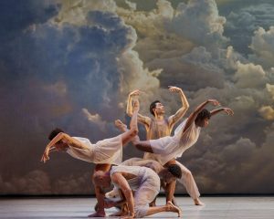 Company of Dancers from the Alvin Ailey Dance Theatre Company performing Many Angels at Sadlers Wells theatre. They are wearing neutral tone clothes with a vibrant could backdrop in the background.