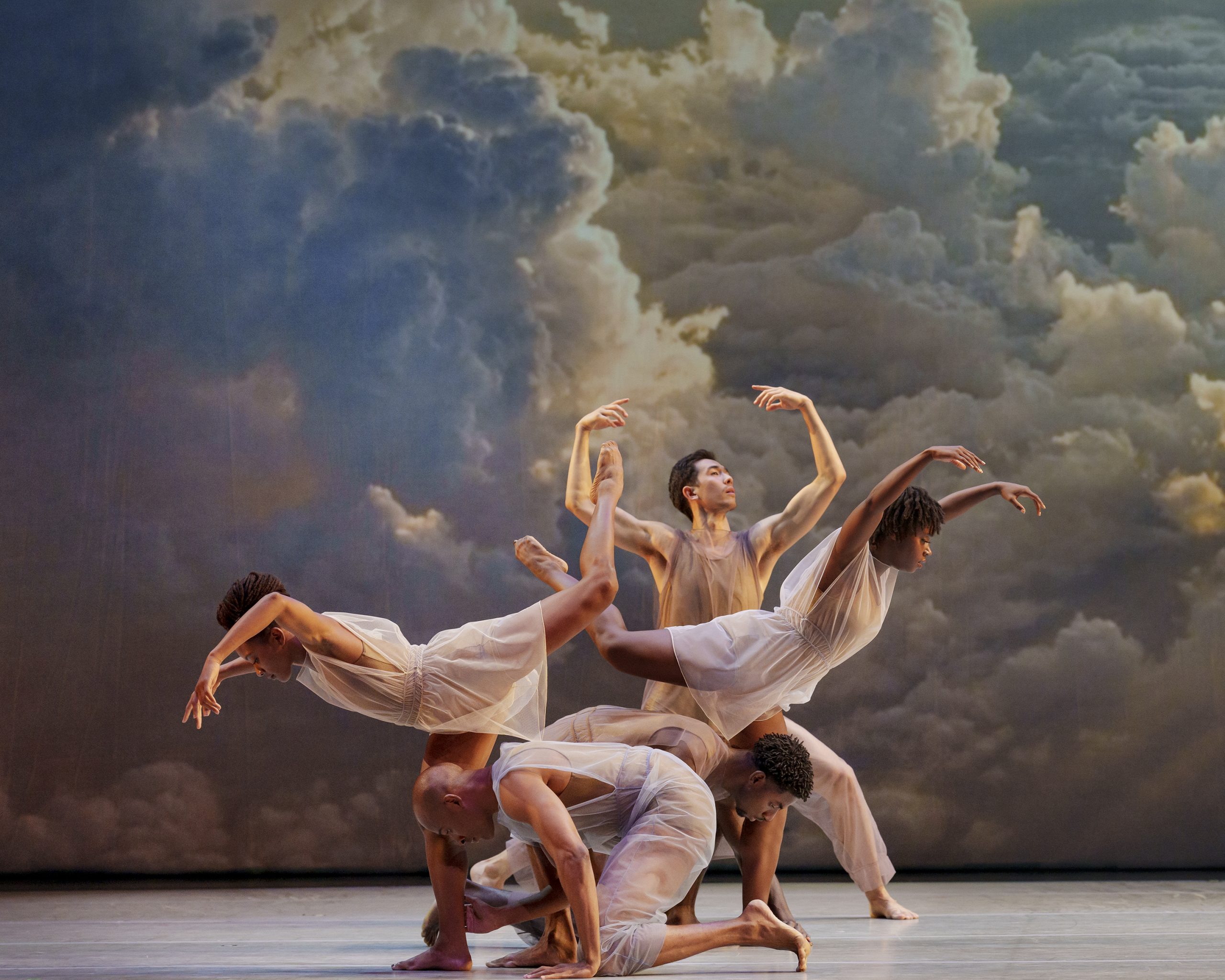 Company of Dancers from the Alvin Ailey Dance Theatre Company performing Many Angels at Sadlers Wells theatre. They are wearing neutral tone clothes with a vibrant could backdrop in the background.