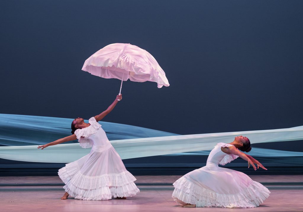 Photograph of two dancers from the Alvin Ailey Dance Theater dressed in white. One is holding an umbrella
