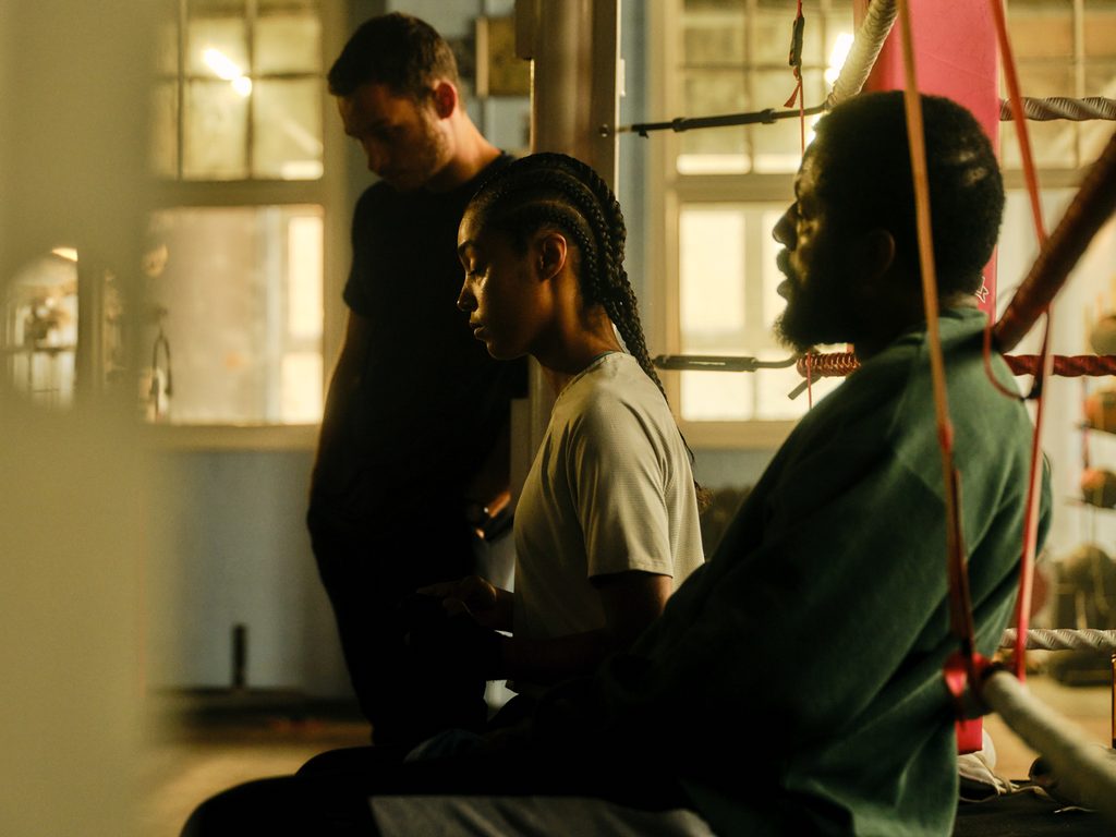 Photograph taken in a boxing ring. A young black woman sits down with cornrows with an older man sat beside her. From film In the Shadows. World Premiering at 70th BFI London Film Festival