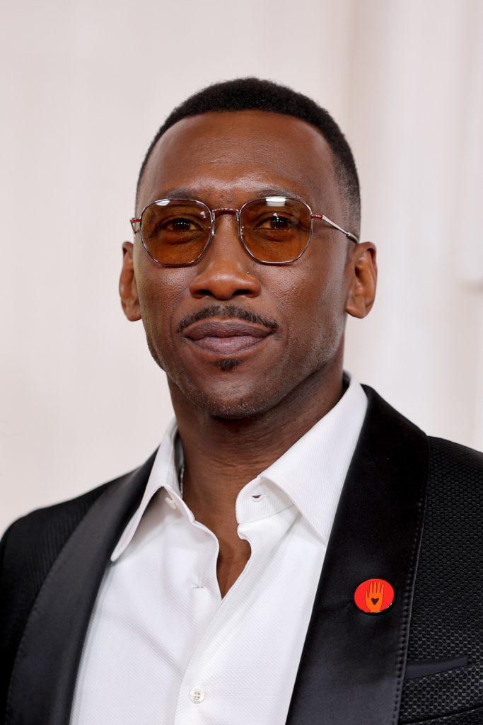 Colour portrait of Actor Mahershala Ali, taken at the 96th Annual Academy Awards in 2024. He is wearing a black tuxedo blazer with an Artists4Ceasefire badge, an unbuttoned white collar shirt and tinted sunglasses.